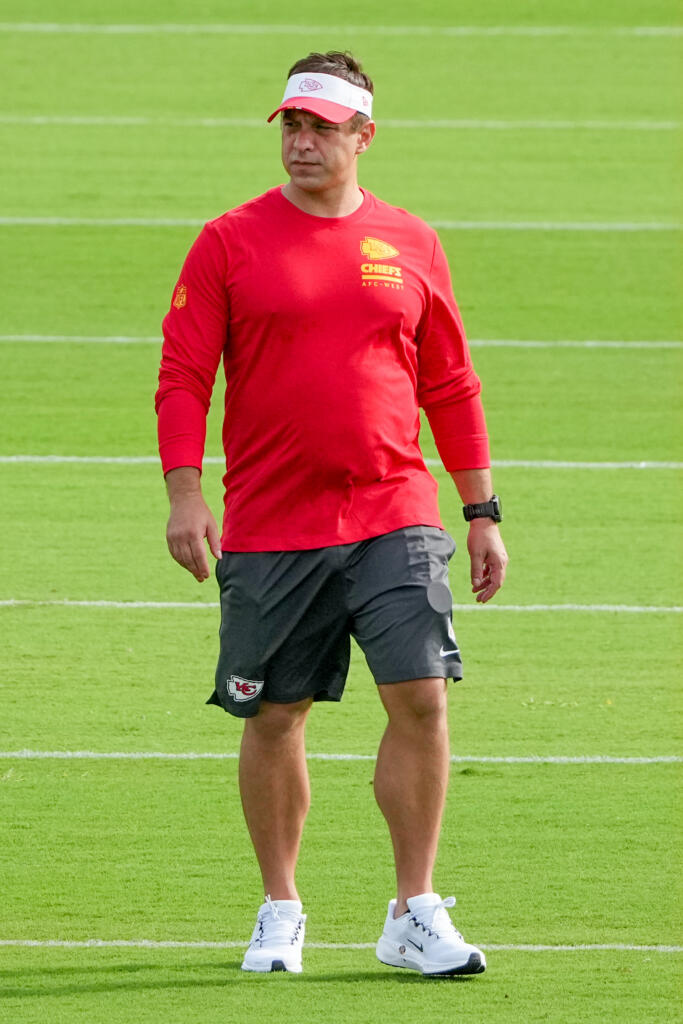 Man in a red Chiefs training shirt and white visor standing on a football practice field with green turf nearby.
