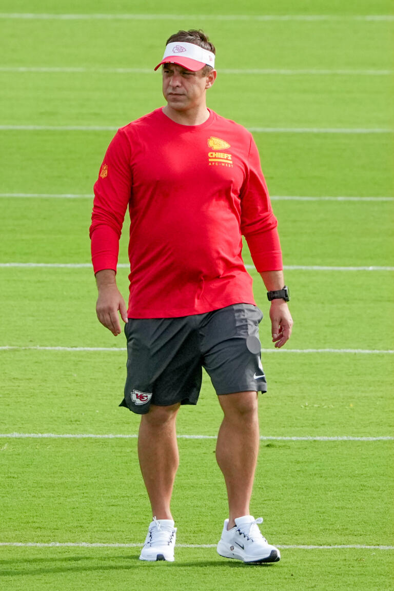 -  Man in a red Chiefs training shirt and white visor standing on a football practice field with green turf nearby.