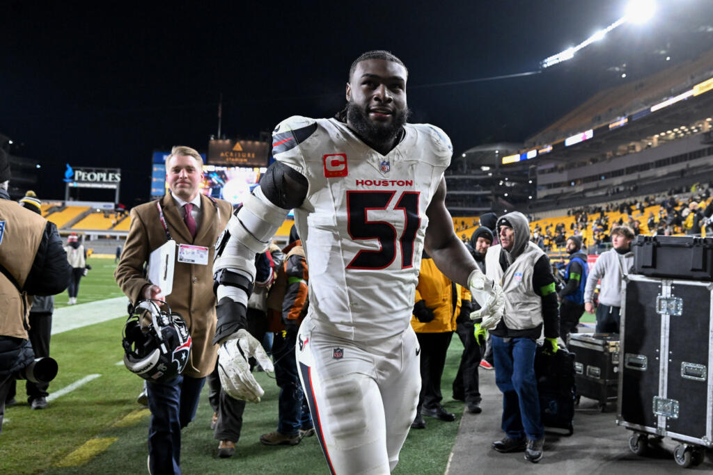 -  Houston Texans player in white jersey #51 walks off the field, helmet in hand, with stadium lights and crowd in the background.