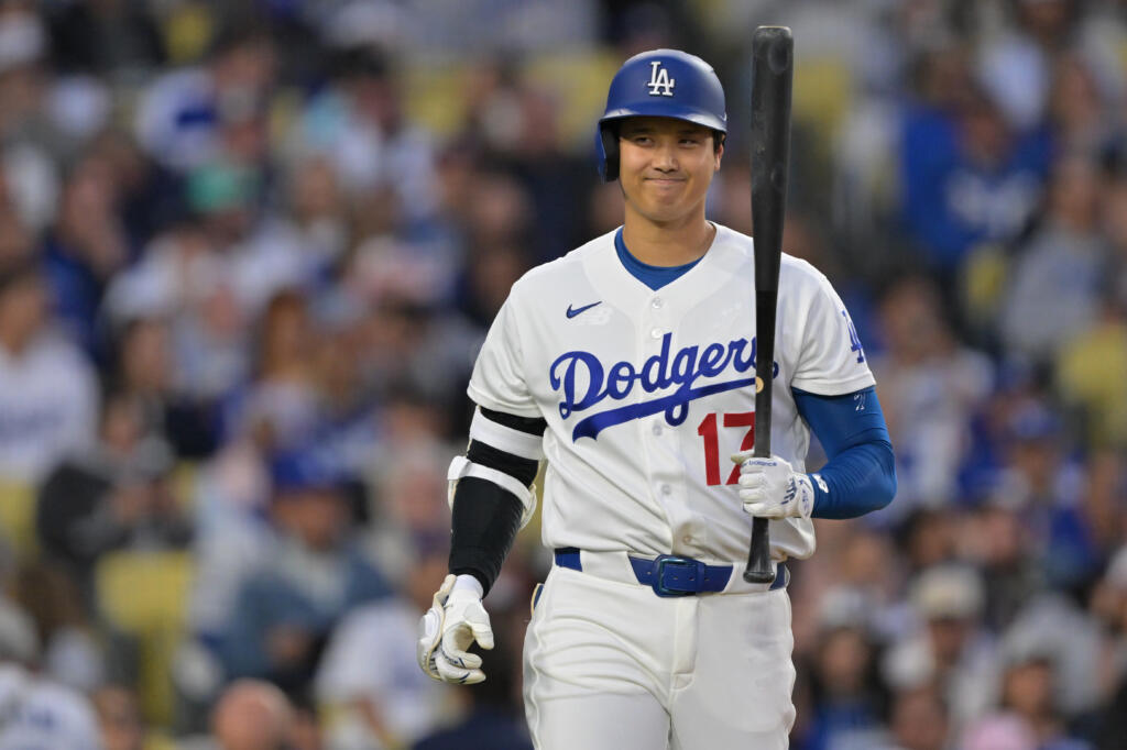 Dodgers player in white uniform with blue lettering, holding a bat and smiling at the stadium, wearing helmet and number 17