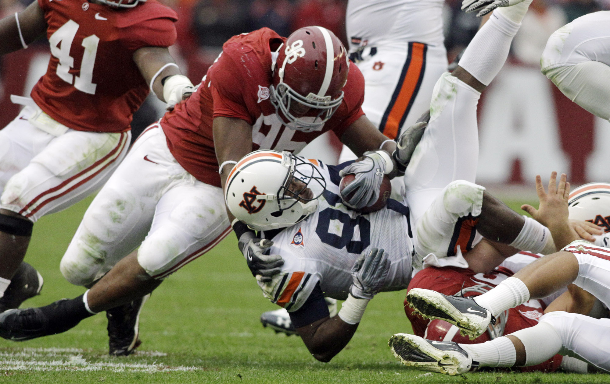 -  Tackle scene during a football game: players in red and white jerseys collide as a ball carrier from the white-helmet team is brought down on the grass field